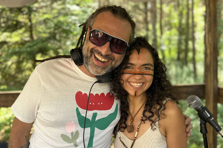 Smiling man with sunglasses and flower print shirt and woman with face paint in a forest in a wooden structure